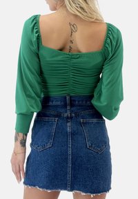 Maglietta verde a maniche lunghe con spalle gonfie e schiena arricciata, abbinata a una minigonna in denim blu aderente con orlo strappato e tasche posteriori.