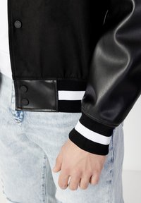 Svart varsityjacka med ullkropp, läderärmar och randiga ribbade muddar. Knappar och en texturerad detalj vid fållen.