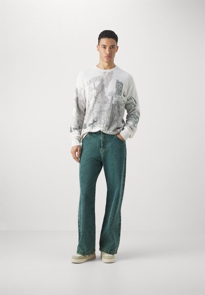 Weißer gerippter Pullover mit grafischen Designs, kombiniert mit lockeren türkisfarbenen Jeans und beigen Schuhen, mit einem entspannten Schnitt und geradem Bein-Design.
