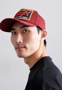 Red baseball cap with a black and yellow patch, featuring "CANADA" and a logo. The cap has a flat brim and a structured shape. Black shirt visible.