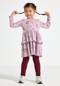 Młoda dziewczynka uśmiechająca się, ubrana w fioletową, kwiecistą sukienkę, burgundowe legginsy, białe tenisówki, trzymająca w górze swoje warkocze obiema rękami.