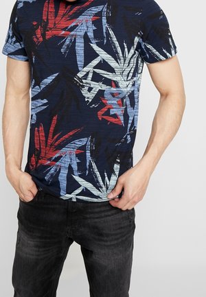 Mężczyzna w granatowej koszulce z czerwonymi, jasnoniebieskimi i białymi abstrakcyjnymi wzorami liści oraz czarnych dżinsach, ręce blisko kieszeni.