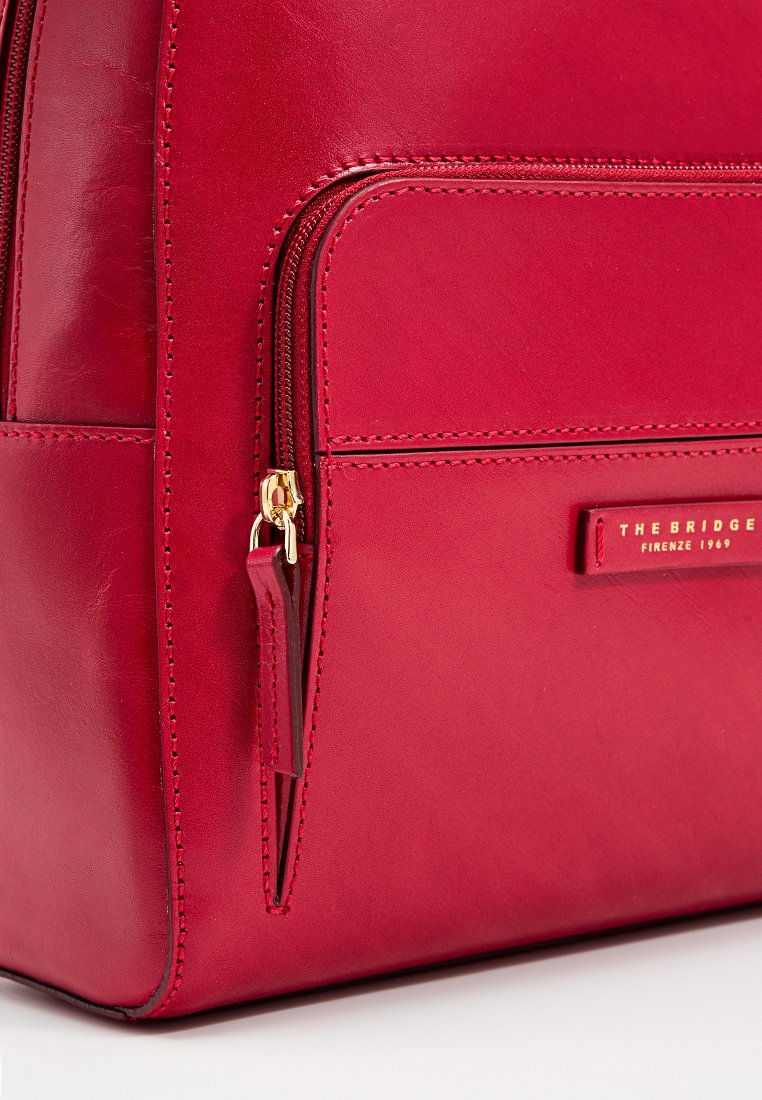 Rote Lederhandtasche mit goldenen Beschlägen, ausgestattet mit einer Reißverschlusstasche, glatter Textur und geprägtem Logo. Kompakt rechteckige Form.