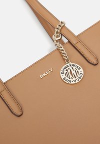 Ljusbrun skinnhandväska med två remmar. Har en guldkedja och en logotypbricka. DKNY-logotyp präglad framtill. Mjuk, strukturerad yta.