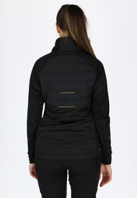 Veste isolante noire avec un col haut, ornée d'accents dorés et d'un logo de marque à l'arrière, fabriquée à partir d'un tissu lisse et texturé.