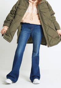 Giubbotto imbottito verde oliva, felpa rosa chiaro con testo bianco e jeans svasati blu scuro. Sneakers bianche completano l'outfit.