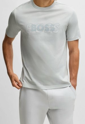 Homme portant un T-shirt gris clair à manches courtes avec le logo "BOSS" et un pantalon gris clair assorti, main ajustant l'ourlet du T-shirt.