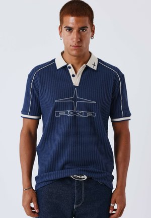 Jeune homme aux cheveux roux courts portant un polo bleu marine texturé avec un col blanc et un logo "PXP", associé à un jean foncé et des accessoires.