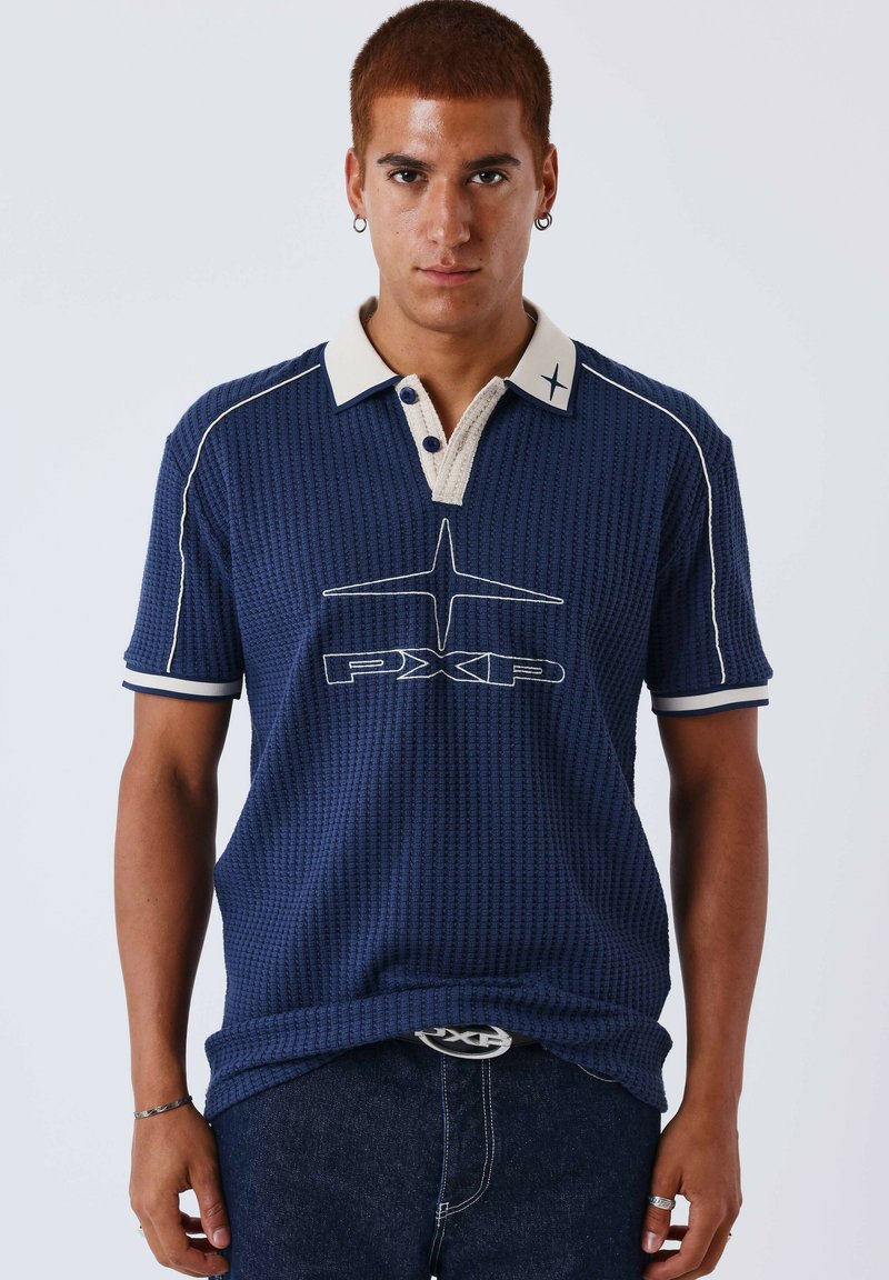 Jeune homme aux cheveux roux courts portant un polo bleu marine texturé avec un col blanc et un logo "PXP", associé à un jean foncé et des accessoires.