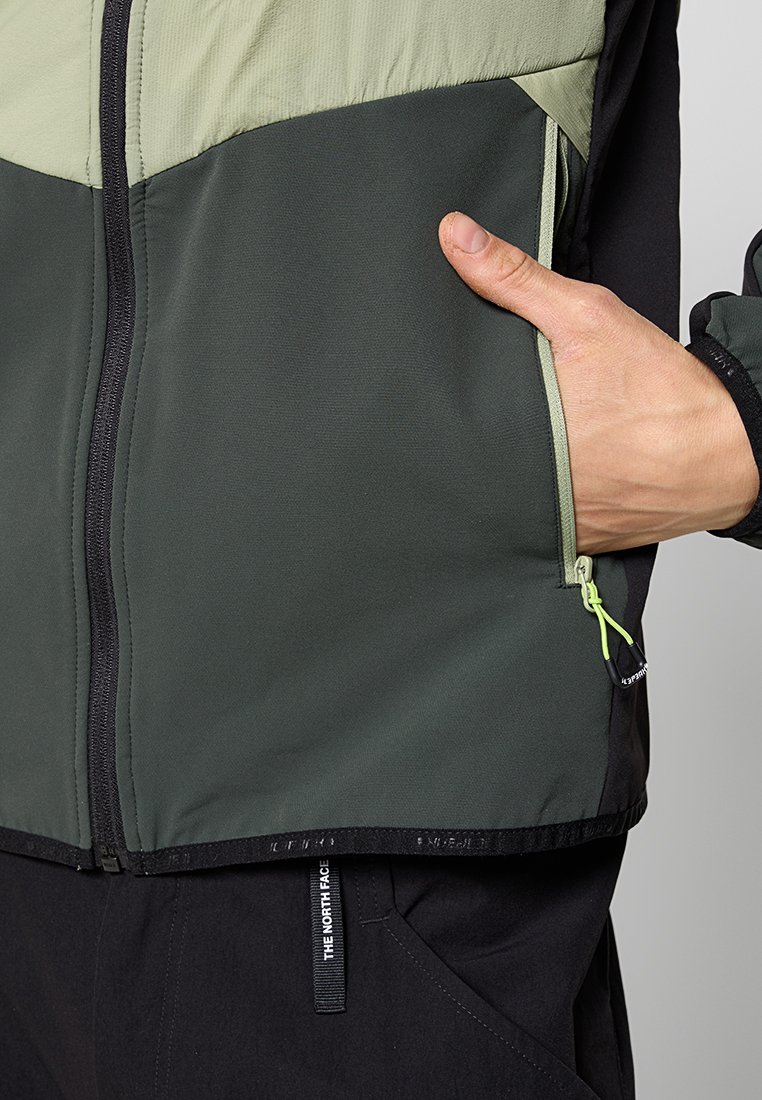Main dans la poche zippée d'une veste bicolore verte et noire portée sur un pantalon noir avec une étiquette "The North Face" visible.
