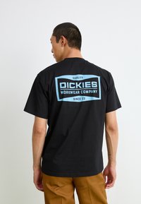 Svart bomullst-skjorte med korte ermer, med stor blå grafikktrykk på baksiden som viser "DICKIES WORKWEAR COMPANY" og "KVALITET."