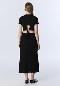 Zwarte jurk met een getextureerd golvend patroon, korte mouwen, een keyhole achterkant en een strik in de taille. Kenmerkt zich door platte, gestifte zwarte schoenen.