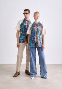 Farverige grafiske t-shirts har en blanding af blå, grøn, rød og gul mønstre. De er kombineret med beige bukser og blå jeans.