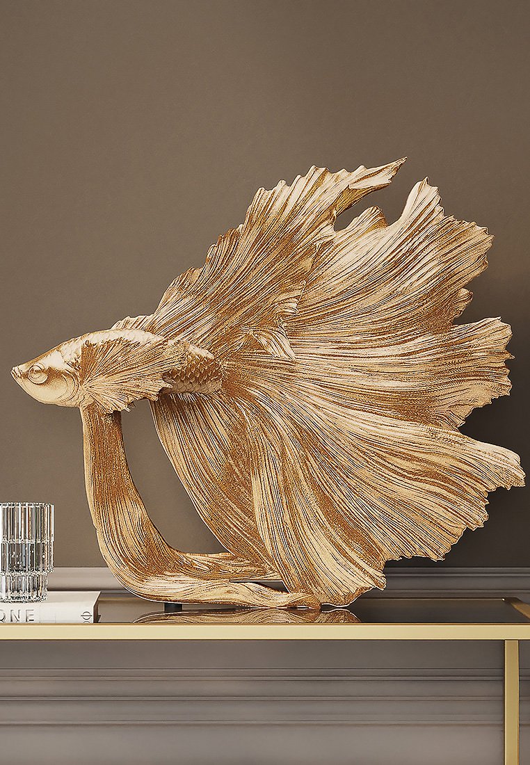 Goldfarbene Fischskulptur mit strukturierten Flossen und einem fließenden Schwanz, detaillierten Schuppen, auf einer flachen Oberfläche neben einem Glasobjekt platziert.