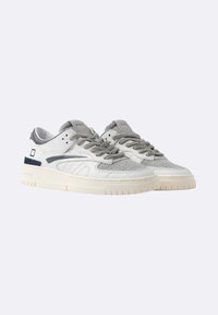 Un paio di sneakers basse bianche e grigie con suole testurizzate, accenti in suede e etichette di marca sulla linguetta su uno sfondo chiaro.