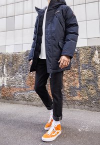 Personne portant une veste à capuche bleu marine, une chemise blanche, un pantalon noir retroussé et des baskets montantes orange vif, se tenant sur un trottoir urbain près d'un mur de graffiti.