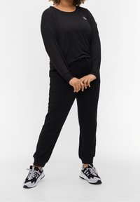 Zwarte lange mouwen top met een ronde halslijn en subtiele logo, gecombineerd met zwarte joggerbroek en zwart-witte sneakers.