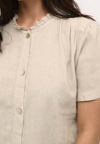 Blusa di lino color beige chiaro con maniche corte, scollatura volant e piccoli bottoni lungo il davanti. Leggera texture visibile sulla superficie del tessuto.