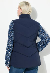 Marinblå puffer väst med hög krage och chevron-quiltat mönster, i kombination med en blå och vit mönstrad ärm.