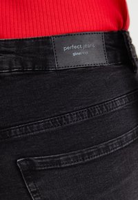 Os jeans de denim preto apresentam uma textura lisa e uma etiqueta com a inscrição "jeans perfeitos" da gina tricot. O design inclui bolsos padrão e detalhes de costura.