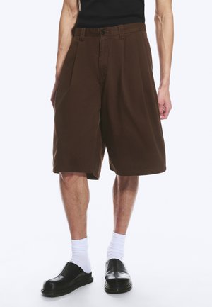 Homme portant un short ample marron foncé jusqu'aux genoux, des chaussettes blanches et des chaussures noires sans lacets sur un fond uni.