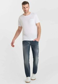 Camiseta blanca de manga corta con un bolsillo texturizado, combinada con pantalones vaqueros de un azul claro y zapatillas blancas con suelas beige. Atuendo simple y casual.