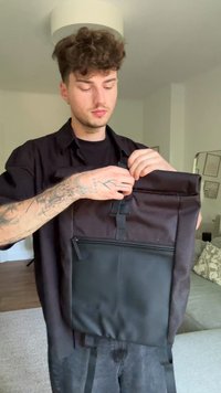 Mochila preta com um tecido texturizado na parte superior e um bolso com fecho ziper em couro liso. Apresenta alças ajustáveis e fecho superior em rolo.