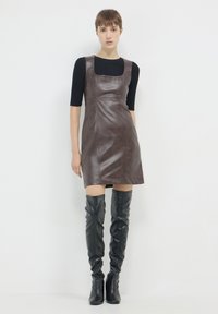 Frau in einem ärmellosen braunen Leder-Minikleid, schwarzem Kurzarmoberteil und schwarzen Overkneestiefeln, die vor einem weißen Hintergrund steht.