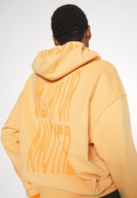 Persona con i capelli ricci corti che indossa un ampio hoodie arancione con grandi testi distorti stampati sulla schiena.