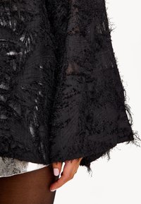 Zwarte blouse met textuur, afgewerkt met rafelige accenten en bloemmotieven, voorzien van een doorschijnende stofdetail en een losse pasvorm.