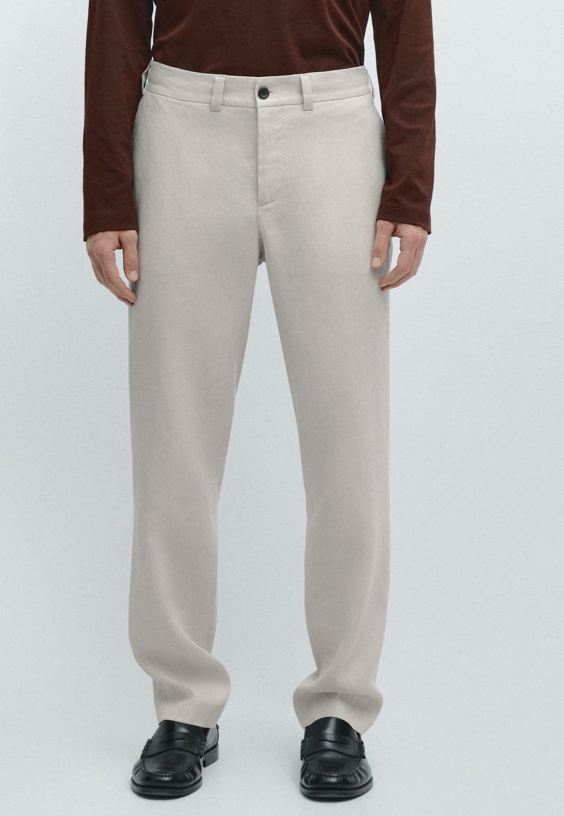 Homme portant un pantalon beige à jambes droites, un haut à manches longues marron et des mocassins en cuir noir, debout devant un fond clair.