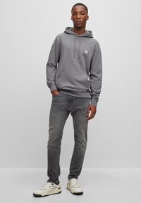 Felpa grigia con cappuccio e cordini, tasca frontale e patch con logo. Abbinata a jeans grigi chiari e sneaker bianche con dettagli neri.