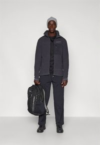 Giacca in pile grigio scuro con cerniera frontale e tasca sul petto. Pantaloni neri, zaino nero con dettagli grigi, cappello di lana grigio, scarpe nere.