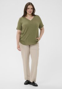 Blouse manches courtes vert olive avec un col en V et une encolure fendue à l'avant, associée à un pantalon large beige clair et des mocassins noirs.