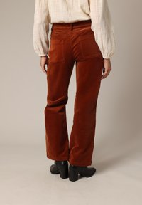 Pantalon en velours côtelé marron avec un design à jambe évasée, doté de deux poches arrière, associé à des bottines à talons noires. Texture lisse visible.