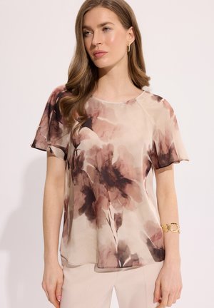 Kobieta o długich falowanych włosach ubrana w krótkiego rękawa, luźną beżową bluzkę z brązowymi kwiatowymi wzorami i jasne spodnie, stojąca na jednolitym tle.