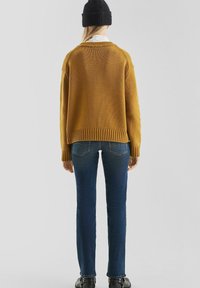 Mosterdkleurige gebreide trui met een geribde zoom en blauwe skinny jeans. De trui heeft een relaxte pasvorm en zichtbare stiksels.