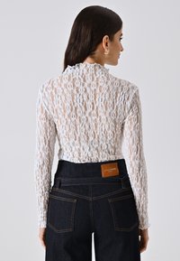 Top bianco in pizzo a maniche lunghe con collo alto e dettagli arricciati, abbinato a jeans in denim scuro a vita alta con un'etichetta in pelle.