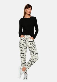 Black ribbed long-sleeve top paired with white pants featuring black tiger stripes. Model wears silver sneakers, hands in pockets.