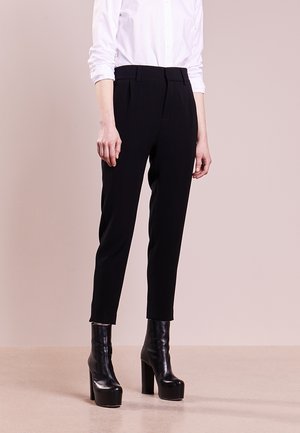 Vrouw draagt zwarte, hooggetailleerde taps toelopende broek, wit shirt met lange mouwen en zwarte enkellaarsjes met plateauhakken, staand op een beige vloer.