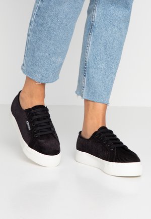 Stopy w czarnych sztruksowych sneakersach na platformie z grubymi białymi podeszwami i czarnymi sznurowadłami, w zestawieniu z jasnoniebieskimi skróconymi dżinsami.