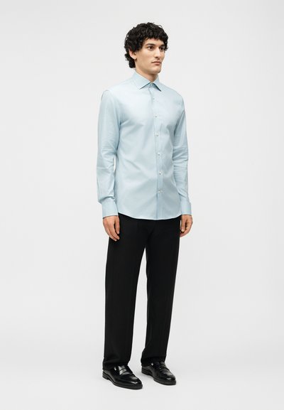 Άνδρας που στέκεται φορώντας ένα ανοιχτό μπλε πουκάμισο με κουμπιά, μαύρο παντελόνι και μαύρα παπούτσια σε απλό λευκό φόντο.