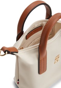Μικρό κρεμ τσάντα χειρός με καφέ δερμάτινες λαβές, χρυσό λογότυπο, εσωτερική τσέπη με φερμουάρ και μεταλλικό δαχτυλίδι στο πλάι.