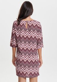 Kobieta z długimi ciemnymi włosami, ubrana w fioletowo-biały sukienkę w zygzakowaty wzór do kolan, z trzema czwarciami rękawów, odwrócona plecami.