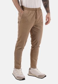 Uomo con pantaloni beige a cordoncino e sneakers bianche, in piedi con una mano in tasca, avambracci tatuati visibili.
