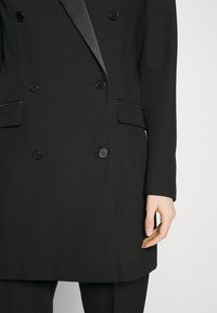 Svart dubbelknäppt kappa med satinéger lapeller och fickor med flik, bärs tillsammans med matchande svarta byxor, där den högra handen hänger avslappnat vid sidan.