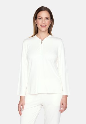 Frau mit glatten braunen Haaren, die ein weißes Langarmshirt mit Vorderreißverschluss und passende weiße Hosen trägt, lächelt in die Kamera.