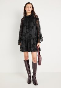 Crna, prozirna haljina s cvjetnim vezom, visokim izrezom, rukavima u obliku zvona i volanima na rubu. Nosiva uz visokootporne, sjajne, burgundske čizme do koljena i usklađenu torbu.