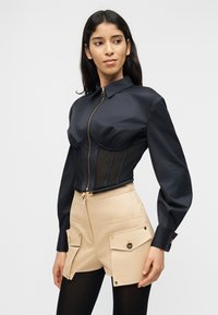 Γυναίκα που φοράει μαύρο τοπ στυλ κορσέ με φερμουάρ και μακριά μανίκια, μαζί με μπεζ σορτς ψηλόμεσα με μεγάλες μπροστινές τσέπες και μαύρο καλσόν.