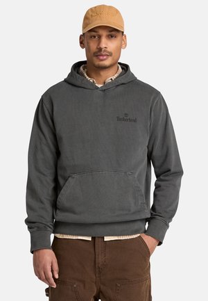 Man draagt een beige Timberland pet, een donkergrijze Timberland hoodie en een bruine broek, staand met één hand in de zak tegen een witte achtergrond.